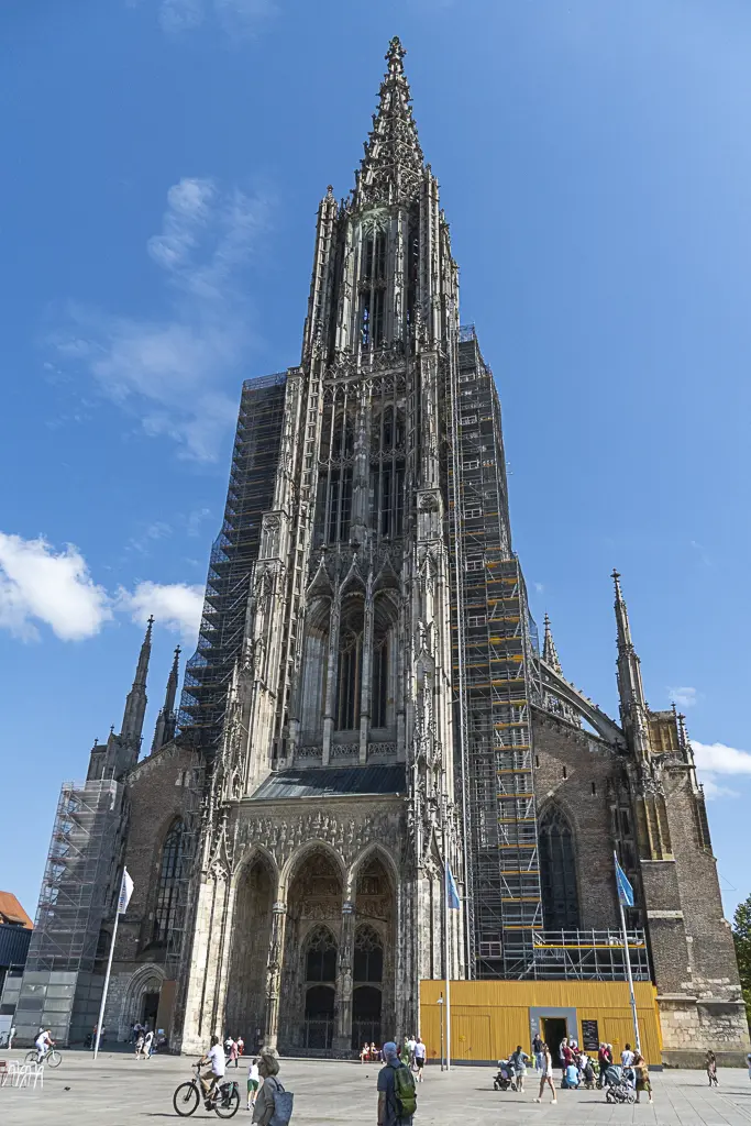 Hier ist ein Alternativtext für dieses Bild:  Eine Totale des gotischen Münsters in Ulm (Ulmer Münster), aufgenommen von der Münsterplatz-Seite. Der zentrale, extrem hohe Kirchturm, der höchste der Welt, dominiert das Bild und ist fast vollständig von Baugerüsten umgeben, was auf laufende Restaurierungsarbeiten hindeutet. Die Fassade besteht aus dunklem Stein und Ziegeln. Im unteren Bereich befindet sich ein gelber Bauzaun oder eine temporäre Struktur, und davor spazieren oder stehen mehrere Menschen, einige mit Fahrrädern, unter einem hellblauen Himmel mit einigen weißen Wolken.