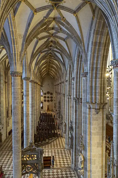 A high-angle interior view of a minster (the Ulm Minster), showing the side aisle and the choir. The scene is characterized by tall, slender, ribbed columns and pointed arches that lead into a cross-ribbed vault. The floor has a striking red-and-white checkerboard pattern. In the depth of the space, rows of dark church pews or choir stalls can be seen. A single person, small compared to the architecture, stands on the floor. The light falls from above, illuminating the light stone columns and the vault.