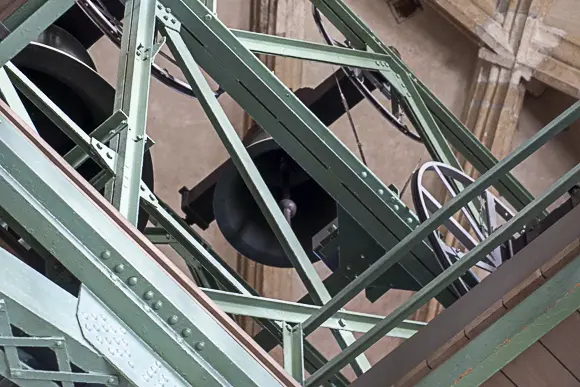 Certainly, here is an alternative text for the image of the bells: A close-up of the interior of a bell tower, showing one or two large, dark church bells. The bells are suspended in a complex and massive framework of green or turquoise painted steel beams, rivets, and braces. You can also see the wheel or suspension used to ring the bells. The background consists of light stone or masonry of the tower.